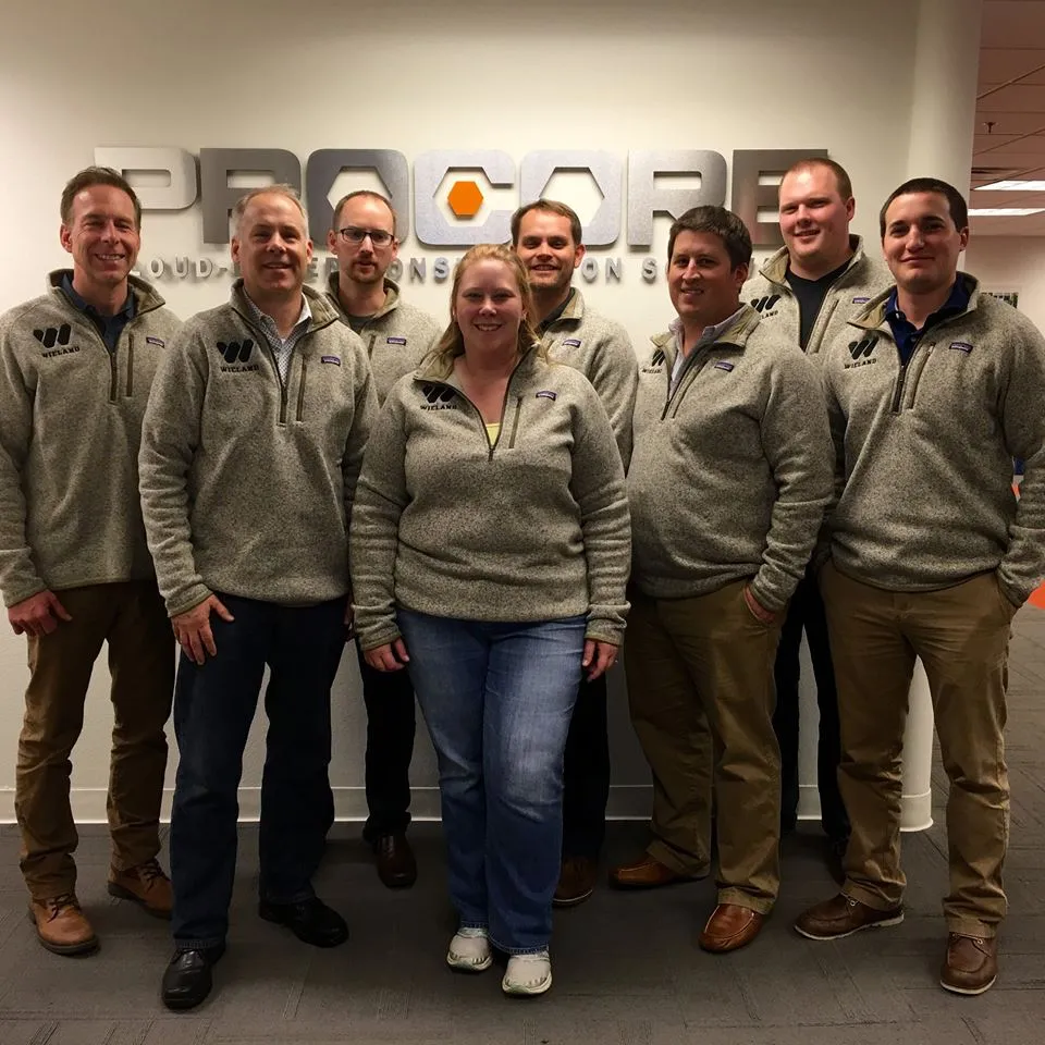 WIELAND team members in matching branded sweaters take a group photo in front of Procore signage