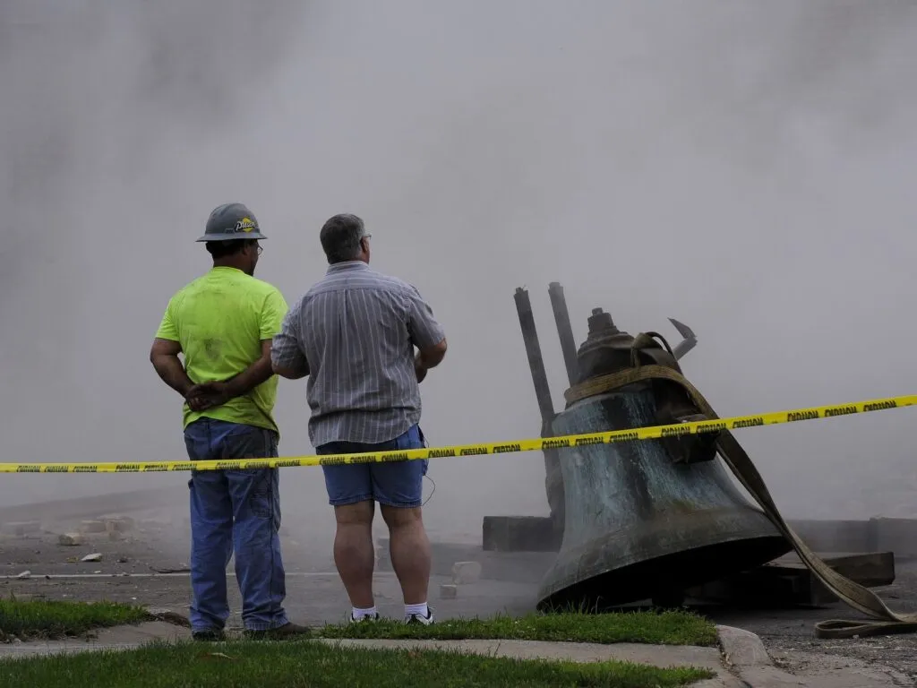 Two men stand side-by-side next to collapsed bell, inside taped off damaged area