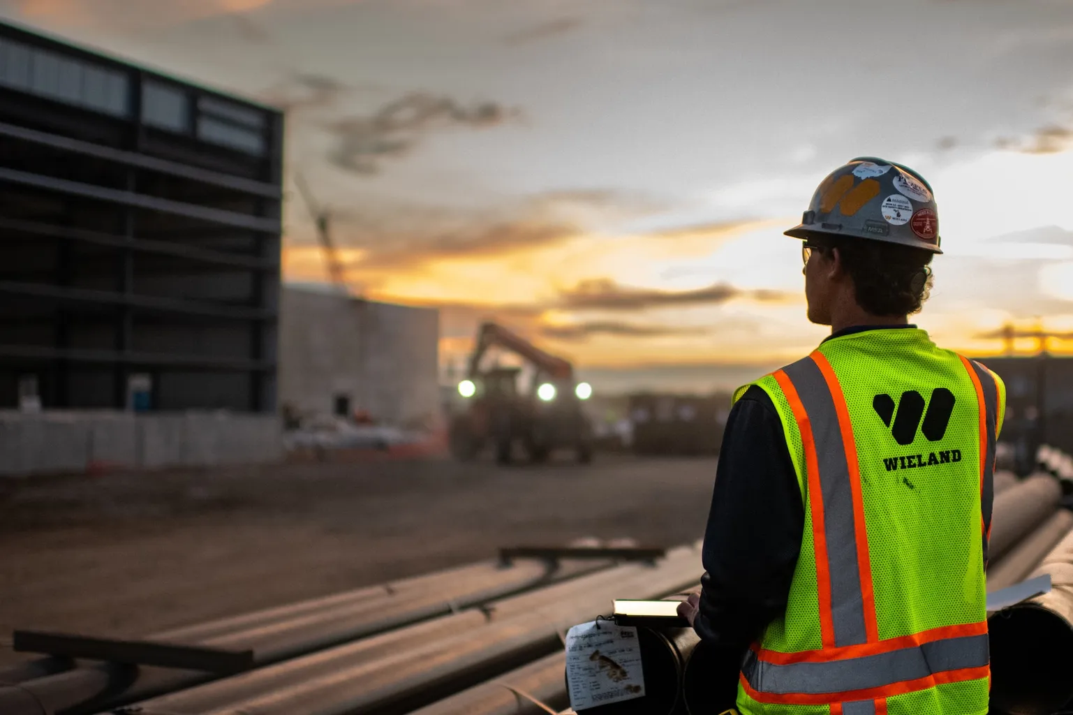 A WIELAND company construction worker wearing a safety vest is silhouetted by a vibrant sunset.