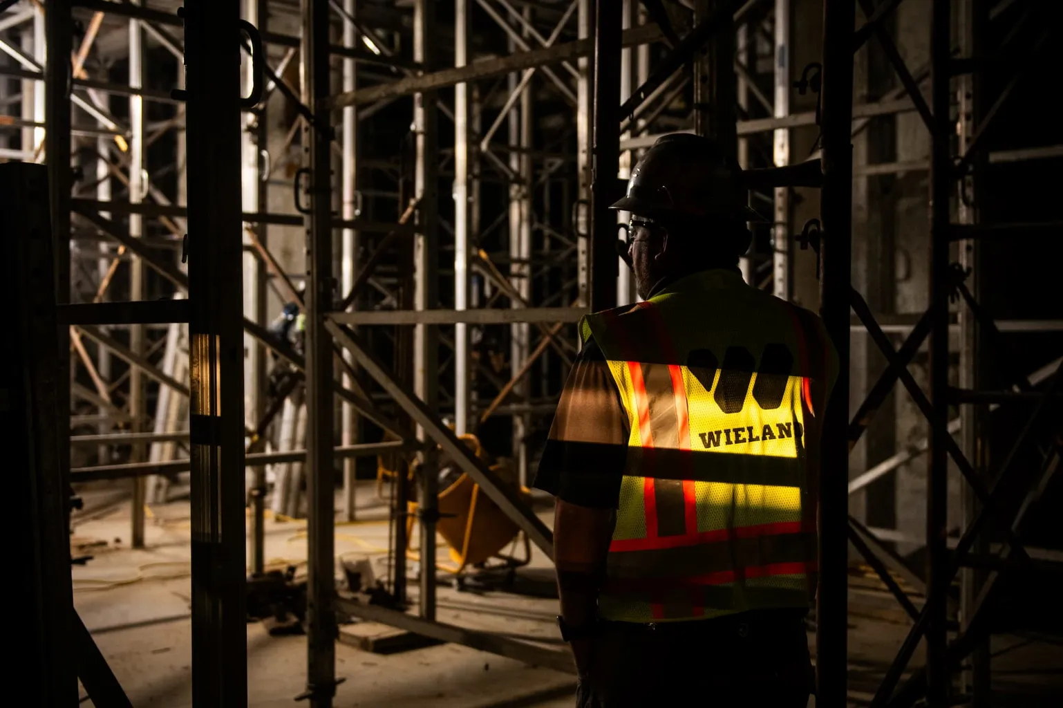 WIELAND employee stands in shadow on a dark job site, with only their safety vest's WIELAND W shining brightly.