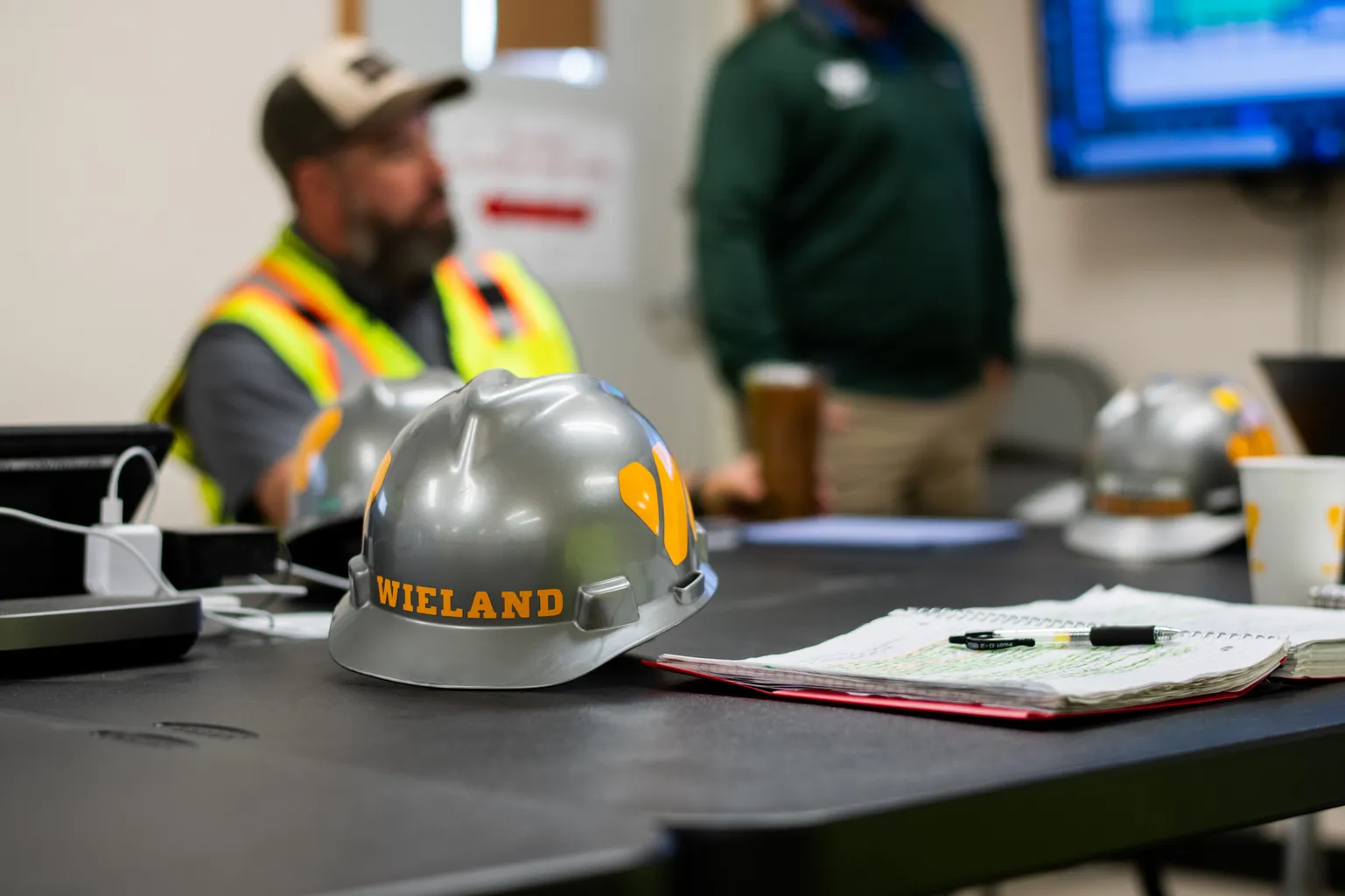 Close up of WIELAND hard hat on table, with employees sat in the background.