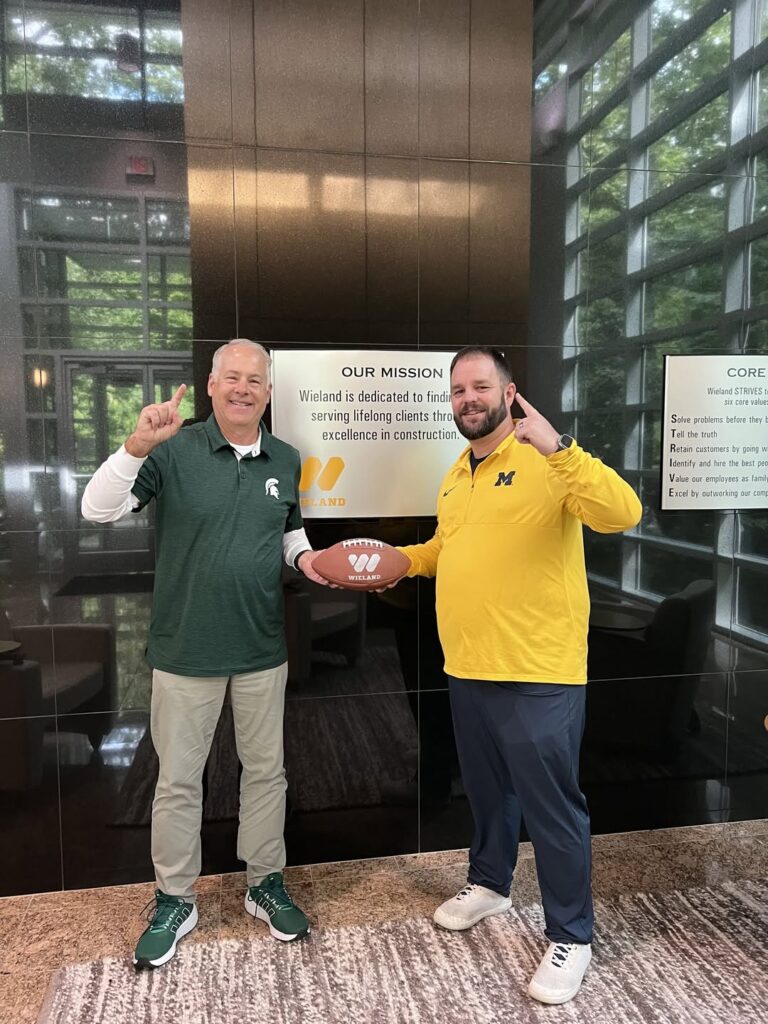 Two men, one wearing Michigan State apparel and the other wearing Michigan gear, holding a football.