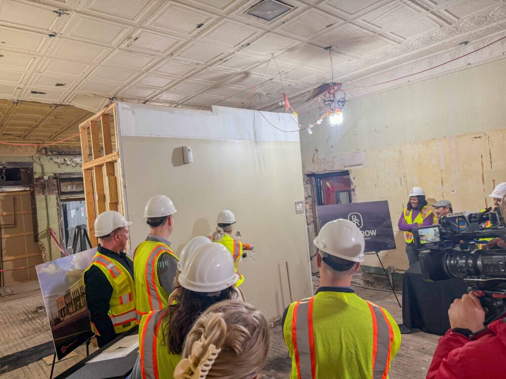 Group in hard hats and safety vests gathered around an employee swinging a hammer at a wall.